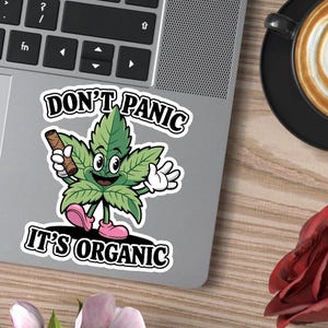 Könnte beinhalten: Ein Aufkleber mit einer Cartoon-Cannabisblatt-Figur, die einen gerollten Gegenstand hält. Das Blatt ist grün mit einem lächelnden Gesicht, trägt rosa Schuhe und weiße Handschuhe. Der Text auf dem Aufkleber lautet "DON'T PANIC IT'S ORGANIC."
