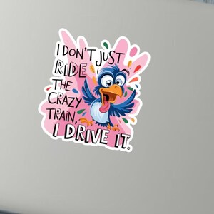 Könnte beinhalten: Aufkleber mit einem Cartoon-Vogel in Blau, Orange und Rosa, vor einem rosa Spritzer-Design. Der Text lautet: "I DON'T JUST RIDE THE CRAZY TRAIN, I DRIVE IT."