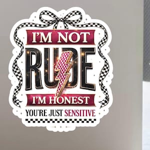 Puede incluir: Una pegatina con el texto "I'M NOT RUDE I'M HONEST YOU'RE JUST SENSITIVE". El diseño presenta una fuente negra y dorada, un rayo rosa y una cinta a cuadros en blanco y negro.