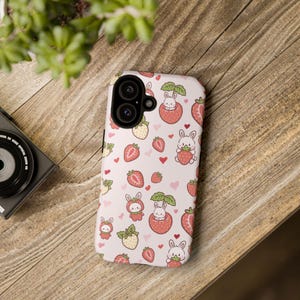 Könnte beinhalten: Eine hellrosa Handyhülle mit einem sich wiederholenden Muster aus Erdbeeren, Herzen und Cartoon-Hasen. Die Hasen sind in verschiedenen Posen dargestellt, einige halten Erdbeeren. Die Hülle hat einen Ausschnitt für eine Dual-Objektiv-Kamera.