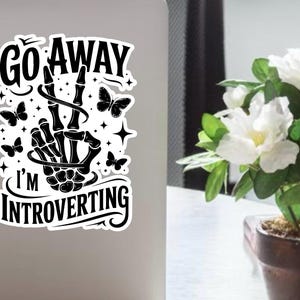 Könnte beinhalten: Schwarz-weißer Aufkleber mit dem Text "Go Away I'm Introverting" und einer Skeletthand, die ein Friedenszeichen zeigt. Das Design umfasst Schmetterlinge und Sterne. Der Aufkleber befindet sich auf einem Laptop.