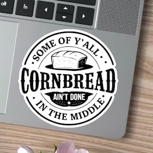 Puede incluir: Una pegatina redonda en blanco y negro con el texto "SOME OF Y'ALL CORNBREAD AIN'T DONE IN THE MIDDLE" que rodea una ilustración de pan de maíz. La pegatina está sobre un portátil, con una taza de café y una rosa en el fondo.