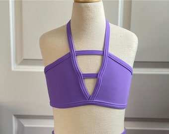 May include: Purple halter top with a criss-cross design in the front. The top has a V-shaped neckline and is made of a smooth, stretchy fabric.