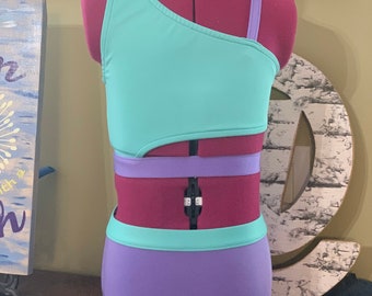 May include: A two-piece swimsuit in teal and purple displayed on a mannequin. The top is a one-shoulder design with a teal bodice and a purple strap. The bottoms are high-waisted and purple. The swimsuit has a cut-out design.