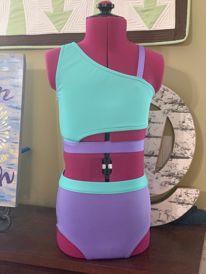 May include: A teal and purple two-piece swimsuit on a mannequin. The top is a one-shoulder design with a teal bodice and a purple strap. The high-waisted bottoms are purple. The swimsuit features a cut-out design.