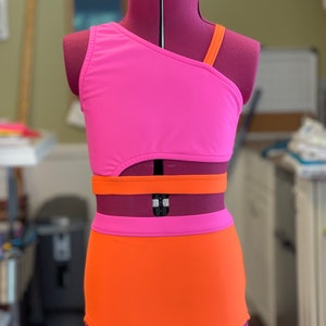 May include: A vibrant two-piece swimsuit in pink and orange, presented on a mannequin. The top features an asymmetrical design with one shoulder strap. The high-waisted bottoms are solid orange. The swimsuit is suitable for swimming or water sports.