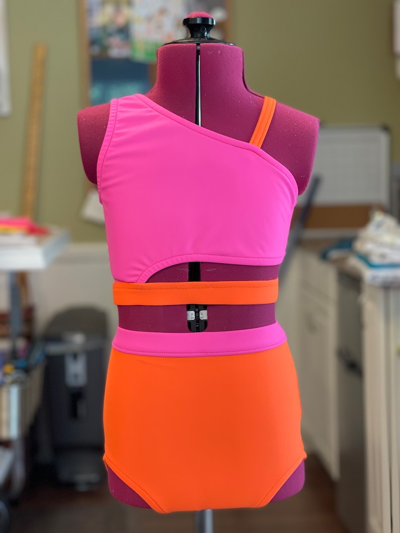 May include: A vibrant two-piece swimsuit in pink and orange, presented on a mannequin. The top features an asymmetrical design with one shoulder strap. The high-waisted bottoms are solid orange. The swimsuit is suitable for swimming or water sports.