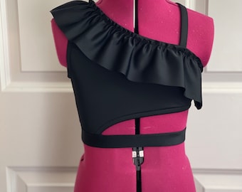 Könnte beinhalten: Schwarzes One-Shoulder-Crop-Top mit Rüschenausschnitt und Cutout-Design. Das Oberteil wird auf einer rosa Schneiderpuppe präsentiert. Das Kleidungsstück hat einen Riemen über einer Schulter und ein Band um die Taille.