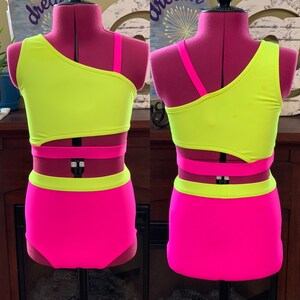 May include: A vibrant dance costume in neon yellow and hot pink, displayed on a mannequin. The top is a one-shoulder design with a cutout, and the high-waisted bottoms are solid pink. The costume has pink straps and accents.