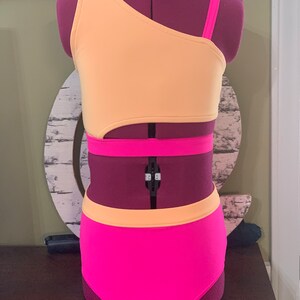 May include: A two-piece dance costume in peach and hot pink. The top is asymmetrical with a single strap and a cutout. The high-waisted bottoms are hot pink with a peach waistband. The costume is displayed on a purple mannequin.