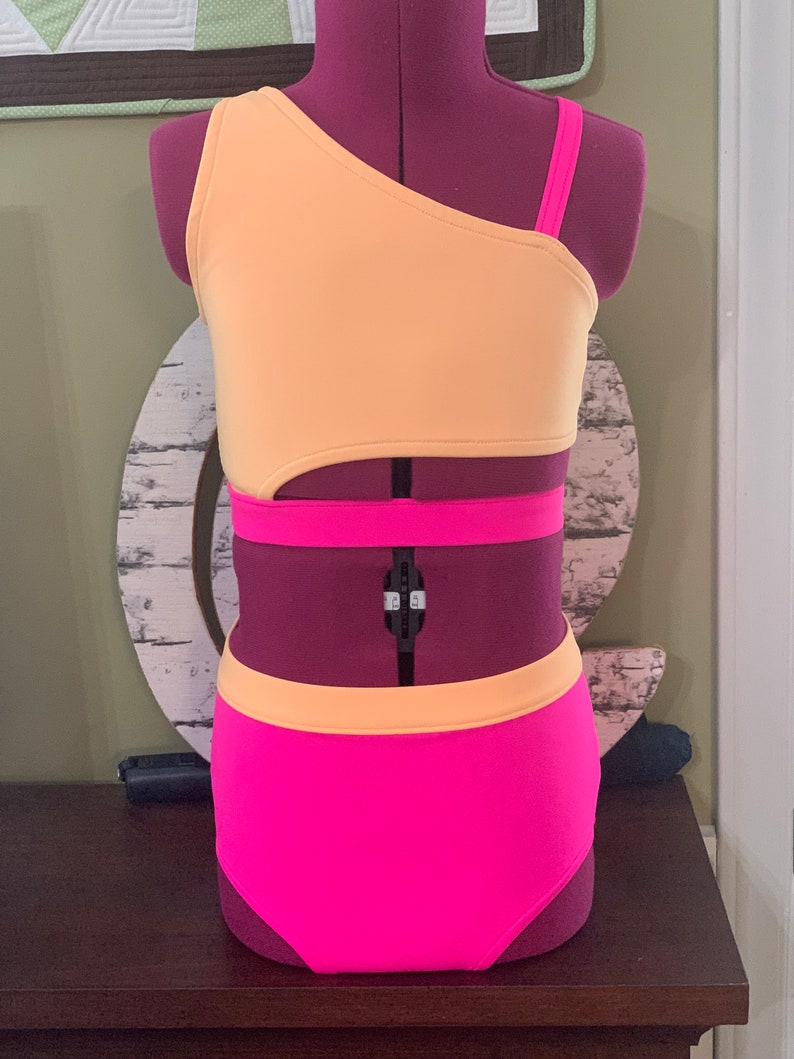 May include: A two-piece dance costume in peach and hot pink. The top is asymmetrical with a single strap and a cutout. The high-waisted bottoms are hot pink with a peach waistband. The costume is displayed on a purple mannequin.