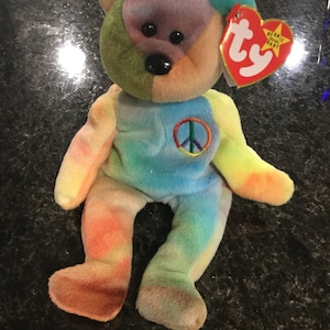 Può includere: Un orsacchiotto Beanie Baby multicolore con motivo tie-dye e un simbolo di pace sul petto. L'orsacchiotto ha un'etichetta Ty rossa con una stella bianca e le parole "Beanie Babies" stampate su di essa.