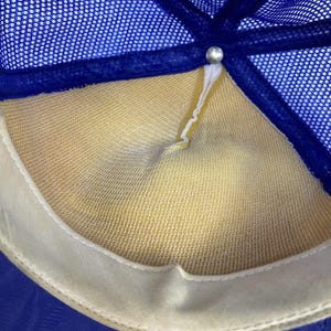 May include: Close-up of the inside of a blue and yellow trucker hat. The hat features a blue mesh top, a yellow interior, and a white sweatband. A silver button secures the hat's panels.