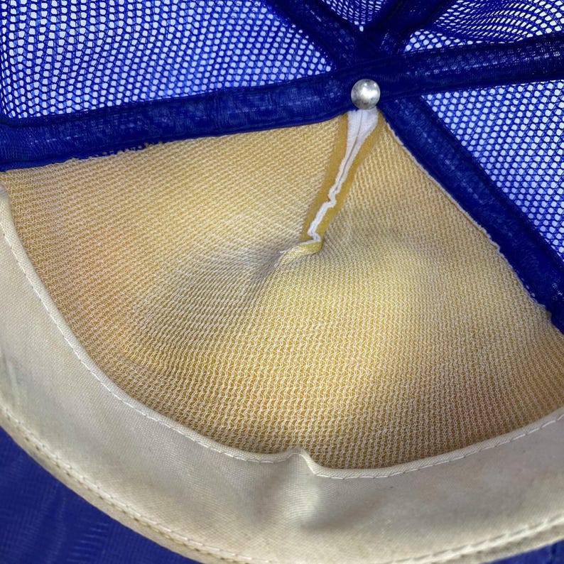 May include: Close-up of the inside of a blue and yellow trucker hat. The hat features a blue mesh top, a yellow interior, and a white sweatband. A silver button secures the hat's panels.