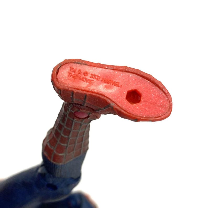 May include: Close-up of a red and blue Spider-Man action figure's foot. The sole of the foot is red with the text "TM & &copy; 2002 MARVEL THE MOVIE". The figure's foot is angled upwards, with a blue and red webbed design.