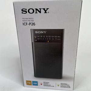 May include: A black Sony ICF-P26 FM/AM radio is shown in its packaging. The radio has a speaker grill and tuning dial. The box features the Sony logo and product information, including FM/AM radio specifications and LED indicators.