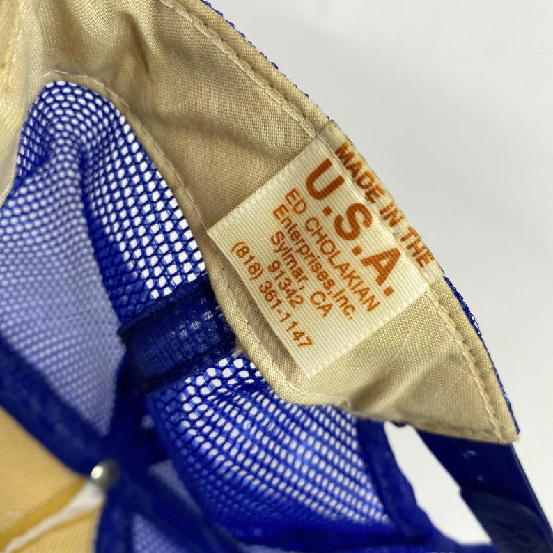 May include: Close-up of a blue mesh trucker hat with a beige interior. The hat features a tag that reads "MADE IN THE U.S.A." with the manufacturer's name and address.