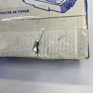 May include: Close-up of a stack of cardboard boxes. The boxes are light gray with blue accents and text. The top box has the words "TOUCHE DE TONER" printed on it. The boxes have some damage, including a tear.