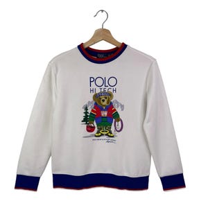 Könnte beinhalten: Weißes Sweatshirt mit Rundhalsausschnitt und langen Ärmeln. Auf der Vorderseite befindet sich eine Grafik eines Bären mit Bandana, Jacke und Stiefeln sowie dem Text "POLO HI TECH". Die Bündchen und der Halsausschnitt haben blaue und rote Verzierungen.
