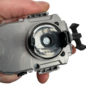 Puede incluir: Un juguete gris y negro con un centro circular y una palanca negra. El juguete tiene el texto "2017 HASBRO SA &copy; TOMY" y "HM, BB, TX VIETNAM" impreso. El juguete est&aacute; sostenido por una mano.