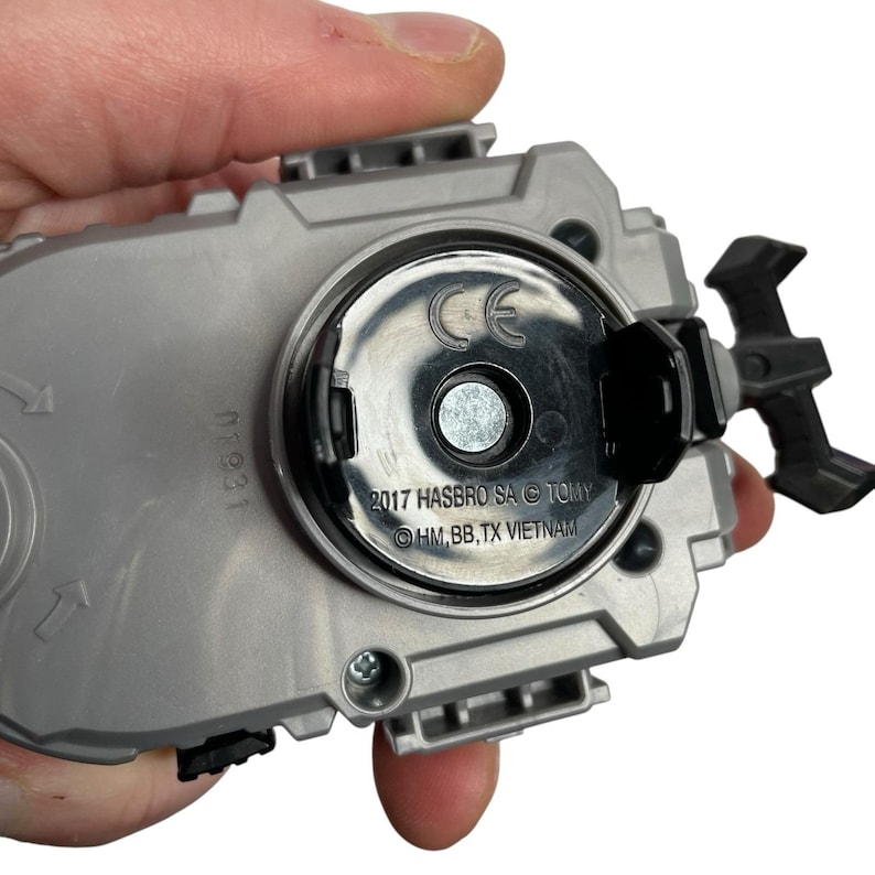 Puede incluir: Un juguete gris y negro con un centro circular y una palanca negra. El juguete tiene el texto "2017 HASBRO SA &copy; TOMY" y "HM, BB, TX VIETNAM" impreso. El juguete est&aacute; sostenido por una mano.