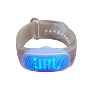 Könnte beinhalten: Ein durchscheinender grauer JBL Fitnesstracker mit blau beleuchtetem Display. Das Display zeigt das JBL-Logo in Blau. Das Armband hat mehrere Verstelllöcher und einen Schlaufenverschluss. Der Tracker ist für das Handgelenk konzipiert.