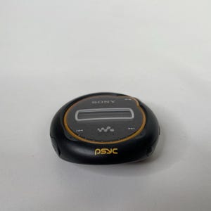 May include: A black and grey Sony digital music player with a circular design. The player has a small screen and control buttons. The word "SONY" is printed on the top, and "psyc" is printed on the bottom in yellow.