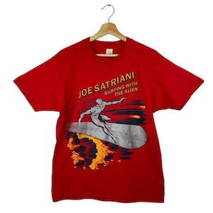 May include: Red t-shirt featuring a graphic of a silver surfer on a surfboard with the text "JOE SATRIANI SURFING WITH THE ALIEN" in yellow and white. The design includes orange and blue accents.