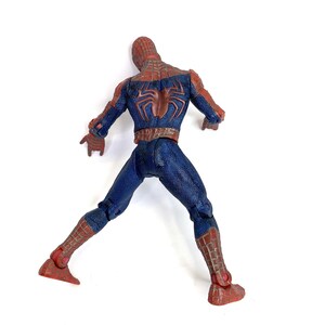 May include: A Spider-Man action figure, viewed from the back, in a dynamic pose. The figure is predominantly blue with red accents, including the mask, forearms, and boots. A large, brown spider emblem is visible on the back.