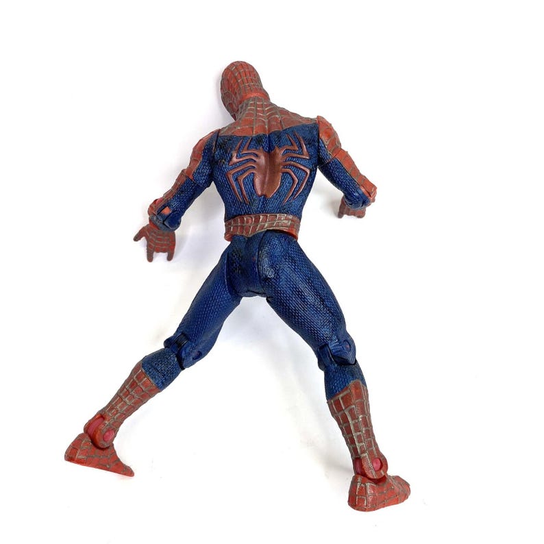 May include: A Spider-Man action figure, viewed from the back, in a dynamic pose. The figure is predominantly blue with red accents, including the mask, forearms, and boots. A large, brown spider emblem is visible on the back.