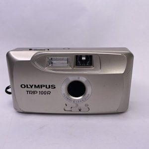 May include: A silver Olympus Trip 100R 35mm film camera. The camera has a built-in flash, a lens with a 27mm focal length, and the Olympus logo. The camera is rectangular with rounded edges and a black strap.