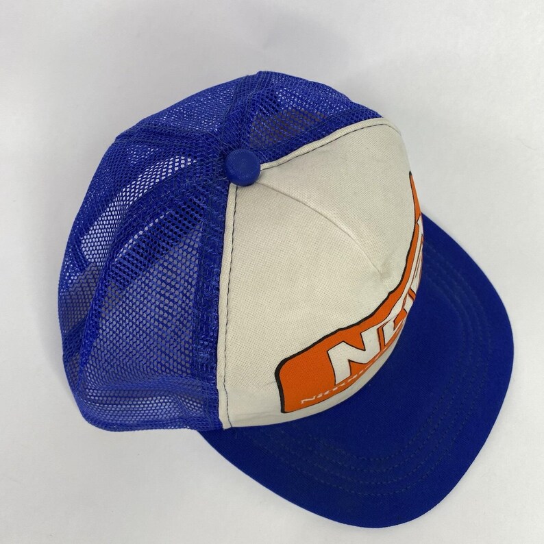 May include: A blue and white trucker hat with a curved brim. The hat features a blue mesh back, a white front panel with orange lettering, and a blue button on top. The hat is a classic style.
