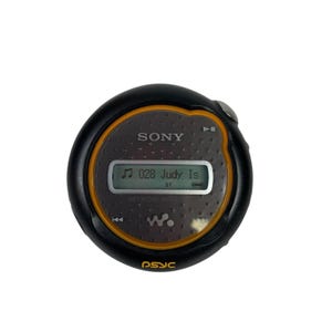 May include: A black and orange Sony Network Walkman MP3 player. The screen displays "028 Judy Is" and "S1". The player has a circular design with a yellow border and the word "psyc" printed on the bottom.
