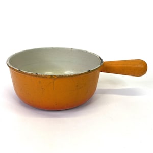 May include: An orange enameled cast iron saucepan with a single handle. The interior is white, and the exterior is a vibrant orange. The pan shows signs of wear, adding to its vintage charm. A classic piece for cooking.