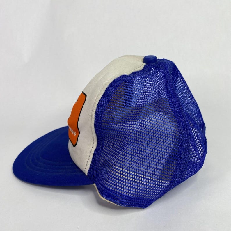 May include: A blue and white trucker hat with a blue brim and mesh back. The hat features an orange patch with white text. The top of the hat is white, and the button is blue.
