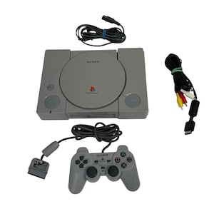 May include: A gray Sony PlayStation 1 video game console with a matching controller, power cable, and AV cables. The console has the PlayStation logo and the word "SONY" printed on it. The controller has the word "SONY" printed on it.