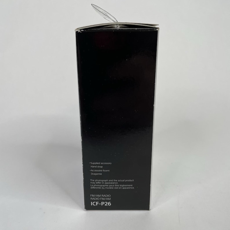 May include: A black rectangular box with the text "Supplied accessory", "Hand strap", "Accessore foun", "Dragonne", "FM/AM RADIO", "ICF-P26". The box has a clear plastic tab extending from the top.