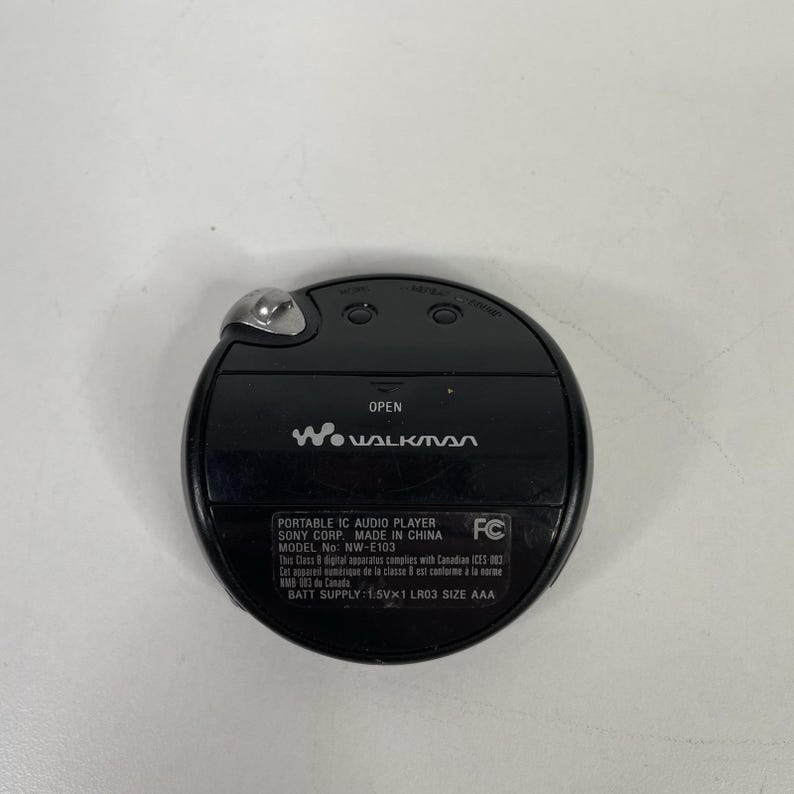 May include: Black Sony Walkman portable IC audio player, model NW-E103. The device has a circular design with a silver button and the Walkman logo. The text "OPEN" is visible above the screen.