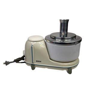 May include: Cream-coloured Bosch food processor with a white bowl and stainless steel accents. The appliance has a rounded design with blue trim and the brand name "BOSCH" is visible. It includes a power cord and plug.