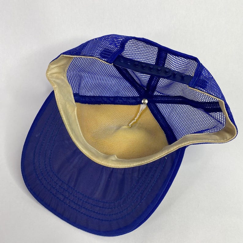 May include: A blue trucker hat with a mesh back and a curved brim. The hat features a snapback closure for adjustable sizing. The interior is lined with a yellow fabric, and the brim is a solid blue color.
