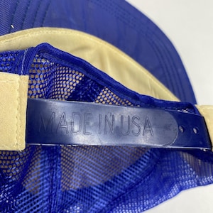 May include: Blue and tan mesh-backed baseball cap with a curved brim. The adjustable strap is navy blue and features the text "MADE IN USA". The cap has a classic trucker hat design.