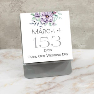 May include: A white card with a floral design in shades of purple and green. The card says "March 4" and "153 Days Until Our Wedding Day".