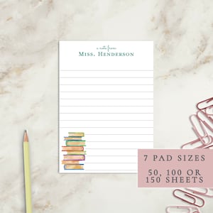 May include: A white notepad with lined paper and a stack of colorful books at the bottom. The notepad has the text "a note from MISS. HENDERSON" at the top. The text at the bottom of the notepad says "7 PAD SIZES 50, 100 OR 150 SHEETS".