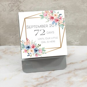 May include: A white card with a gold geometric frame and watercolor floral design. The text reads "September 20, 72 days until our little girl is here."