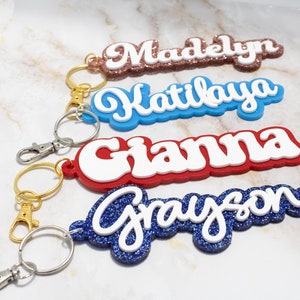May include: Four custom name keychains featuring white script lettering layered over colorful bases. The names Madelyn, Katilaya, Gianna, and Grayson are displayed in glittery rose gold, solid blue, solid red, and glittery blue finishes.