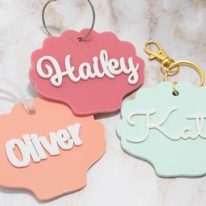 May include: Three personalized luggage tags shaped like seashells. The tags are pink, peach, and light blue. The names "Hailey", "Oliver", and "Katie" are written in white on the tags.