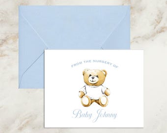 May include: A white rectangular note card featuring a watercolor illustration of a brown teddy bear wearing a white shirt. The text reads FROM THE NURSERY OF and Baby Johnny in light blue. A matching light blue envelope is positioned behind the card.