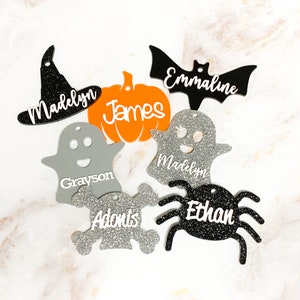May include: A set of seven Halloween-themed ornaments with glitter and names written on them. The ornaments are shaped like a witch's hat, a pumpkin, a ghost, a bat, a spider, a skull and crossbones, and another ghost.