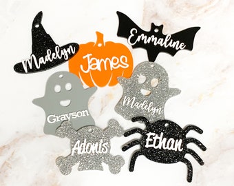 May include: A set of seven Halloween-themed ornaments with glitter and names written on them. The ornaments are shaped like a witch's hat, a pumpkin, a ghost, a bat, a spider, a skull and crossbones, and another ghost.