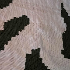 May include: A crocheted blanket with a white background and a dark green design. The design is a geometric pattern of squares and triangles.